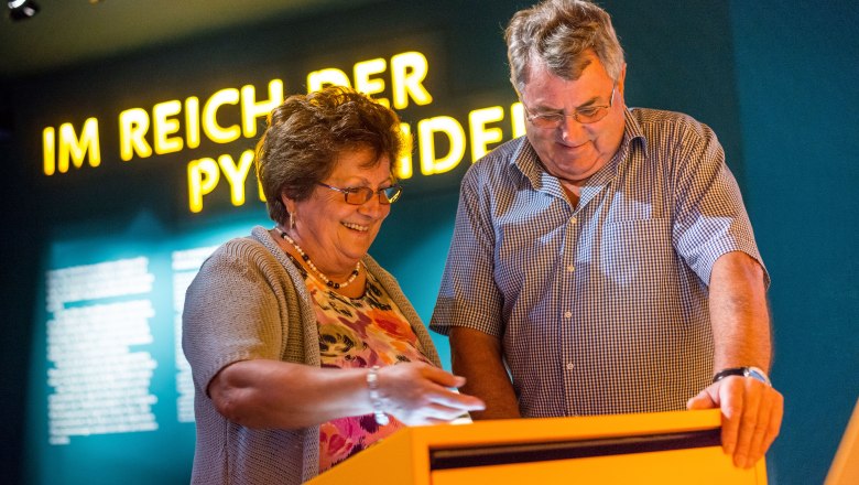 Reise durch die Geschichte, © Sonnenwelt/Klaus Pichler Zwei Personen betrachten ein interaktives Display in einem Museum mit der Aufschrift 'Im Reich der Pyramiden' im Hintergrund.