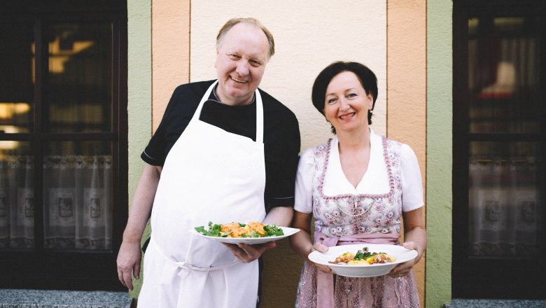 Maria und Alexander Schönauer, © Niederösterreich Werbung/Mara Hohla Ein Mann in Schürze und eine Frau in traditioneller Kleidung halten Teller mit Essen vor einem Gebäude.