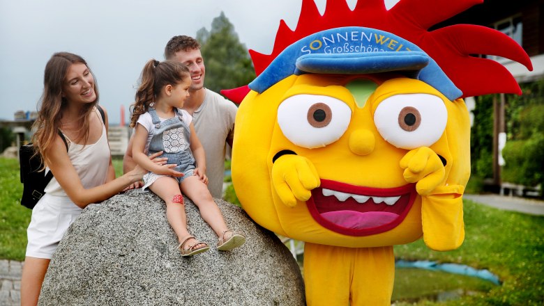 Energie-Erlebnis-Spielplatz, © © Waldviertel Tourismus, weinfranz Eine Familie mit einem Kind sitzt auf einem großen Stein neben einem gelben Maskottchen mit rotem Haar und blauer Mütze im Freien.