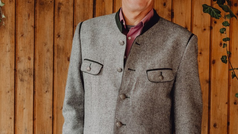 A smiling man in traditional dress in front of a wooden wall with ivy.