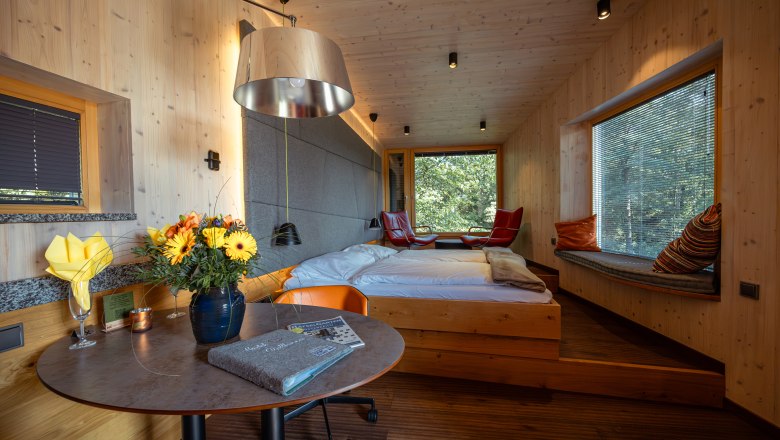 Innenansicht einer modernen Baumhaus-Lodge mit Holzwänden, Bett, Tisch mit Blumen und großen Fenstern mit Blick ins Grüne.