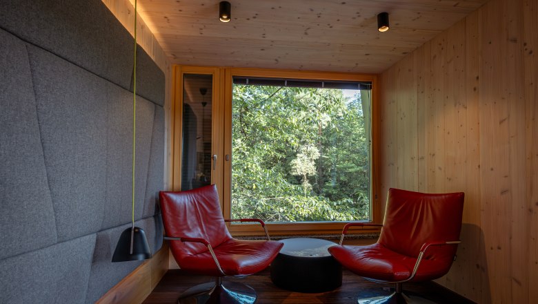 Innenansicht einer modernen Baumhaus-Lodge mit zwei roten Sesseln vor einem großen Fenster mit Waldblick.