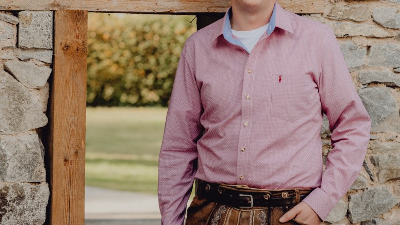Man in traditional leather pants and pink shirt in front of a stone wall.