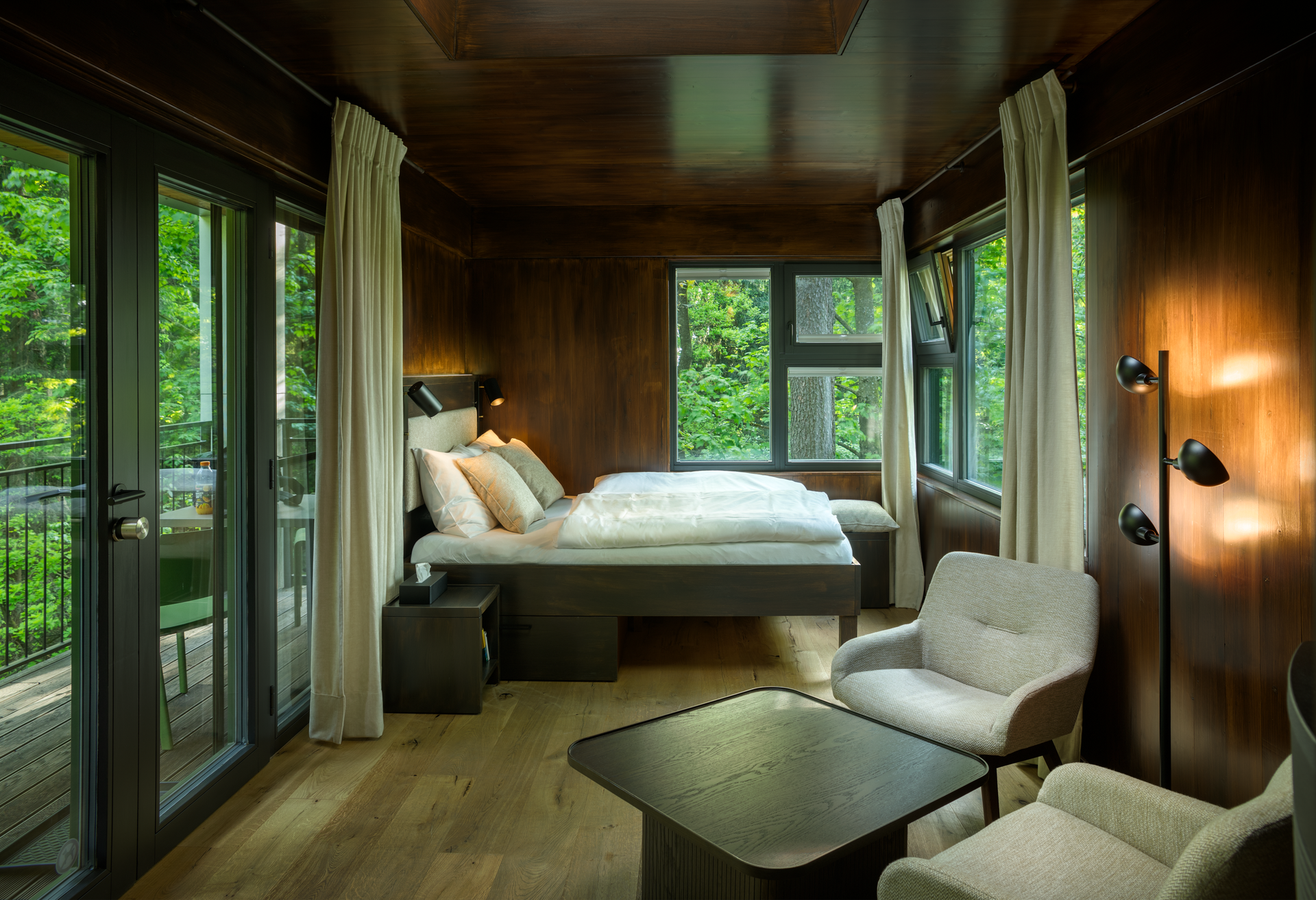 Innenansicht eines modernen Baumhauses mit Bett, Sesseln und großen Fenstern mit Blick ins Grüne.