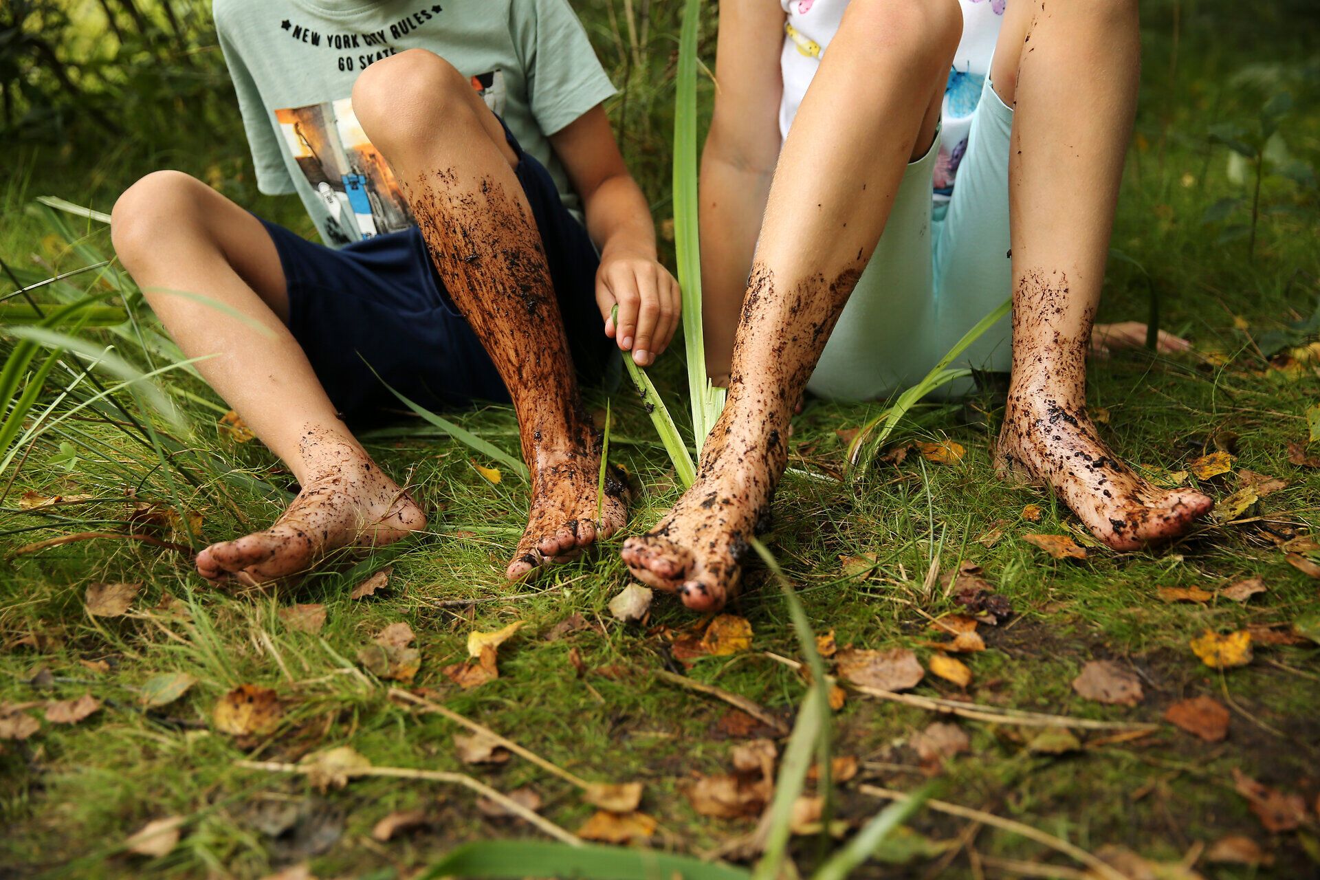 Die Kinder sitzen fröhlich im Hochmoor, ihre Füße sind mit dunklem Moor bedeckt. Umgeben von üppigem Grün und dem sanften Rascheln der Blätter, genießen sie die unbeschwerte Zeit in der Natur.