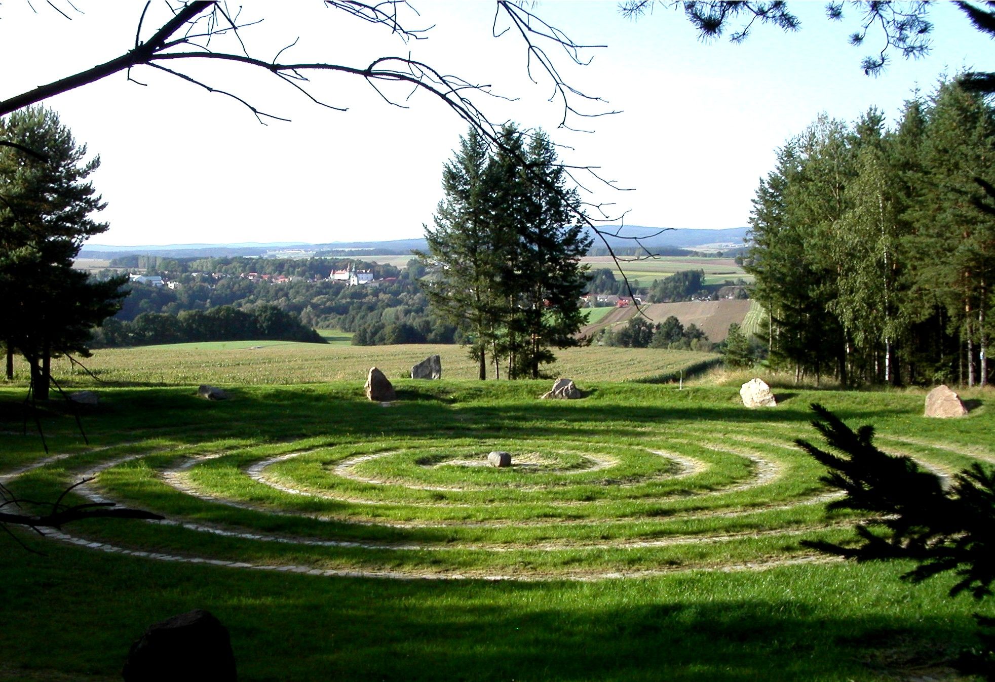 Ein spiralförmiges Labyrinth aus Gras und Steinen auf einer Wiese mit Bäumen und einem Dorf im Hintergrund.