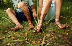 Die Kinder sitzen fröhlich im Hochmoor, ihre Füße sind mit dunklem Moor bedeckt. Umgeben von üppigem Grün und dem sanften Rascheln der Blätter, genießen sie die unbeschwerte Zeit in der Natur.