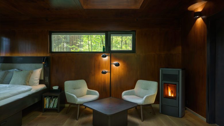 Interior view of a cozy tree house with wooden paneling, two armchairs, a table and a fireplace.