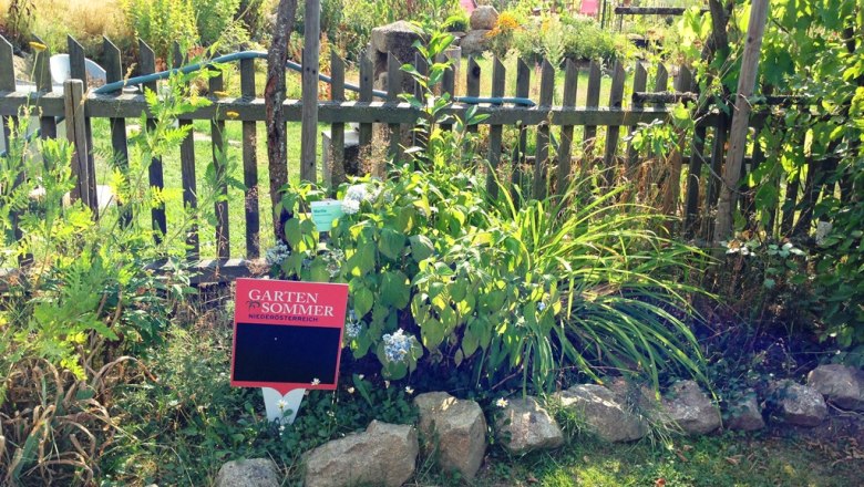 Ein Garten mit Holzzaun, Pflanzen und einem Schild mit der Aufschrift 'Garten Sommer Nieder&ouml;sterreich'.