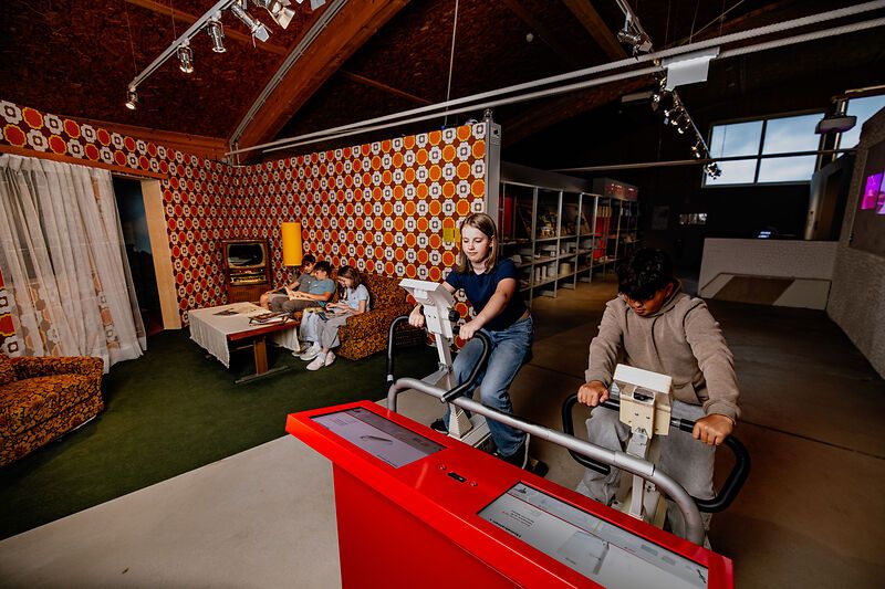 Jugendliche auf Fahrradergometern in einem Raum mit Retro-Wohnzimmer-Dekor.