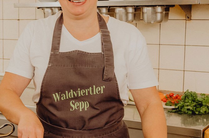 A cook in a kitchen with an apron, wearing "Waldviertler Sepp", stirs in a pan.