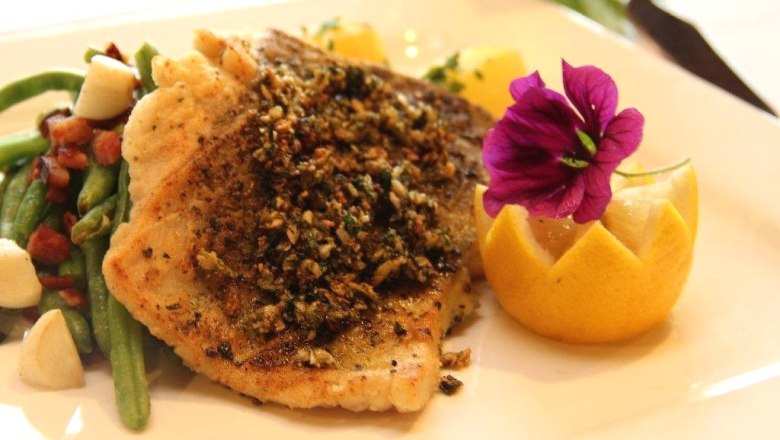 Ein Teller mit gebratenem Fischfilet, dekoriert mit einer Blume und einer Zitronenscheibe, serviert mit gr&uuml;nen Bohnen und Speck.