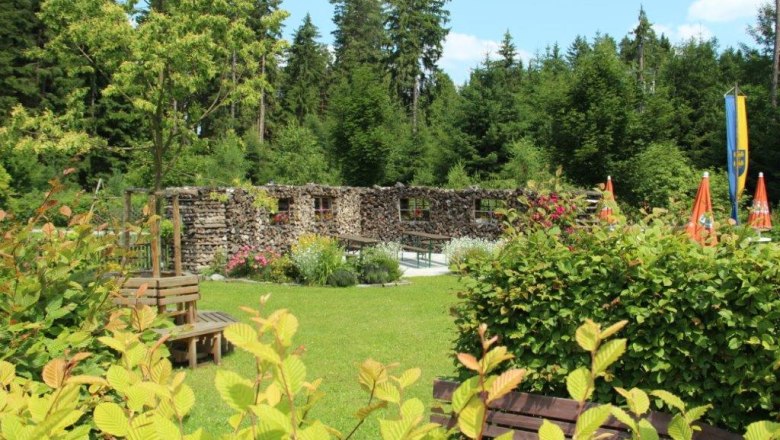 Ein Garten mit Holzwand, B&auml;nken und Sonnenschirmen, umgeben von B&auml;umen.