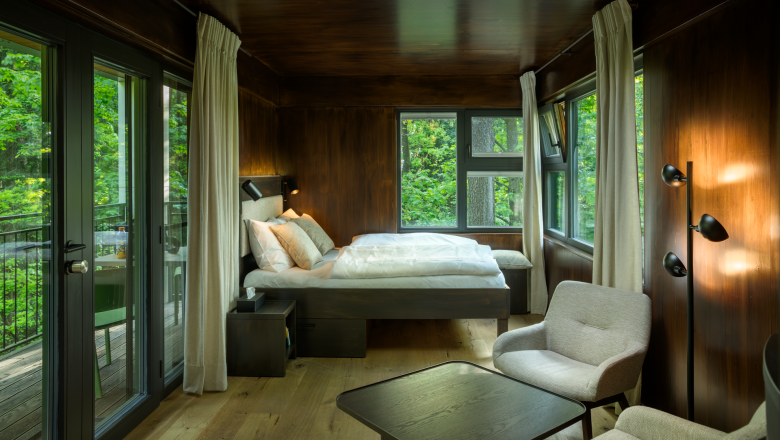 Interior view of a modern tree house with bed, armchairs and large windows with a view of the countryside.