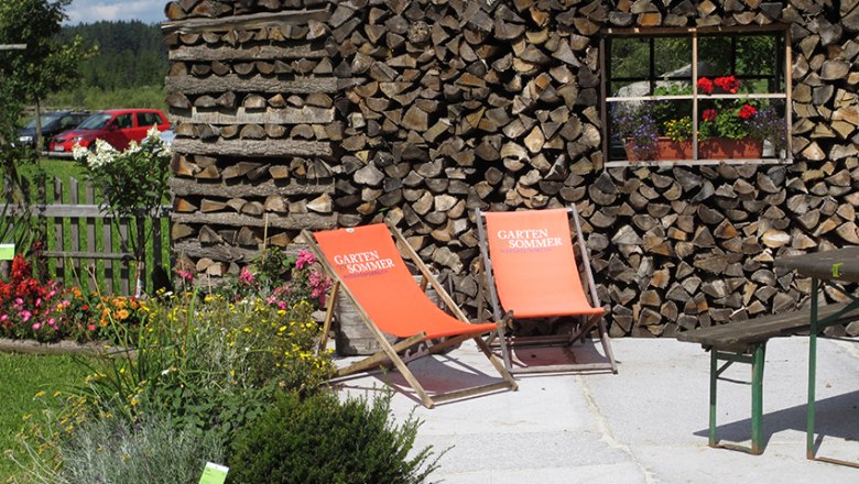 Zwei orange Liegest&uuml;hle vor einer Holzstapelwand im Garten.