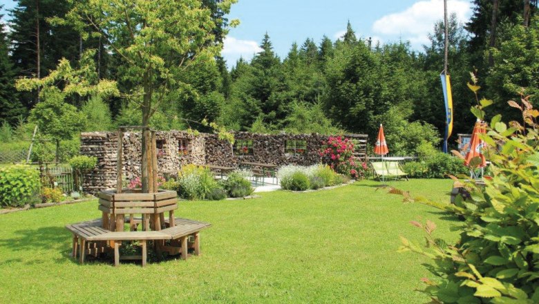 Ein gr&uuml;ner Garten mit Holzb&auml;nken um einen Baum, umgeben von Wald und Blumen.