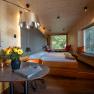 Innenansicht einer modernen Baumhaus-Lodge mit Holzwänden, Bett, Tisch mit Blumen und großen Fenstern mit Blick ins Grüne.